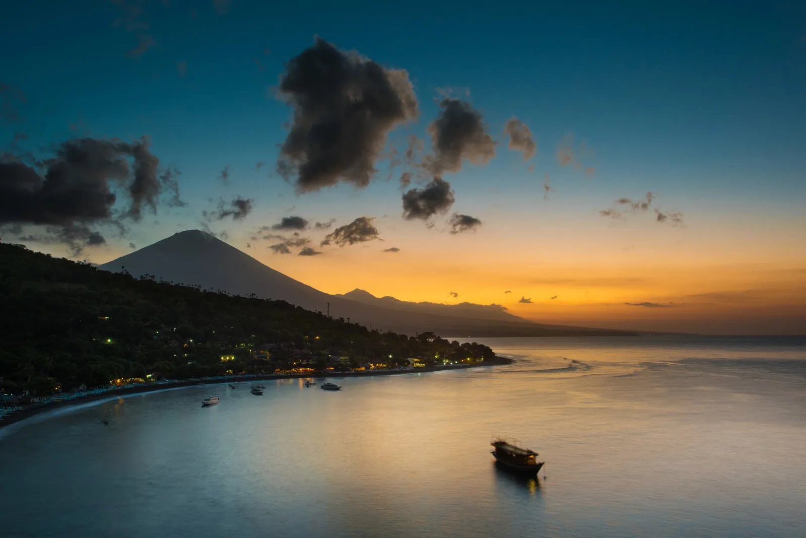 Mount Agung silhouetted against an orange twilight sky above Amed bay, with village lights along the shore and a boat at anchor, Bali