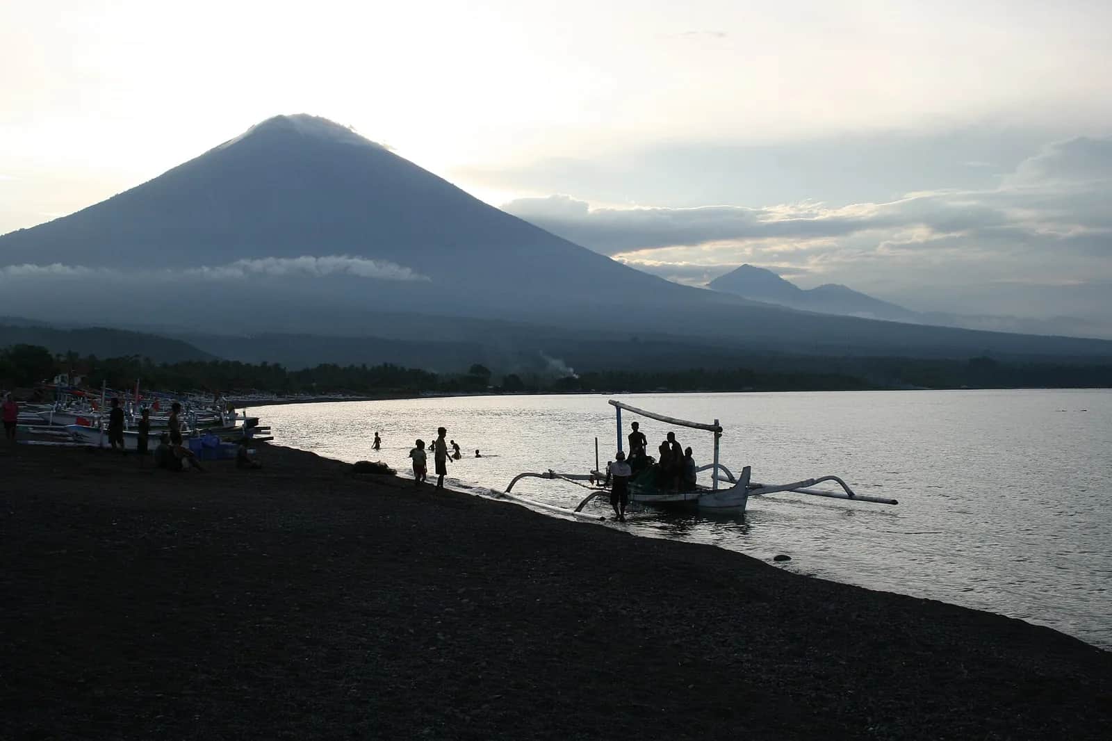 Outrigger jukung boat landing on the black sand of Amed Beach at dusk, with Mount Agung rising behind the bay, east Bali