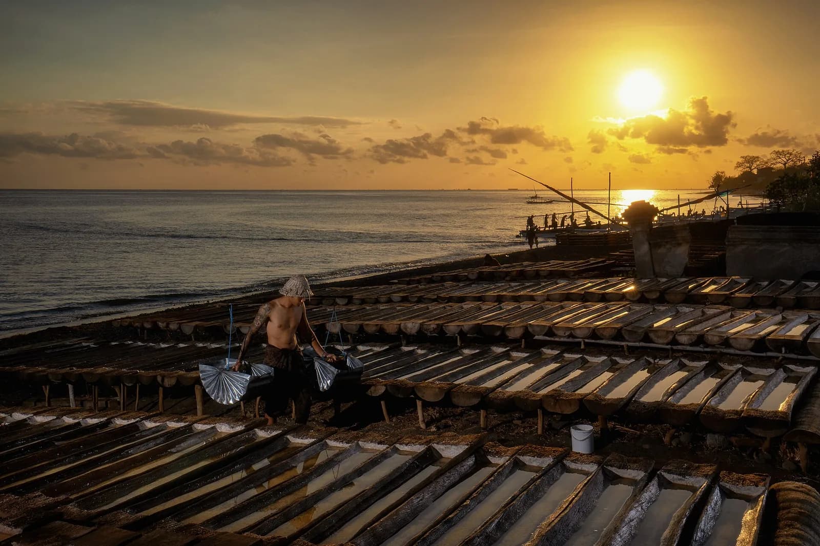 Salt farmer carrying seawater across rows of hollowed palm-trunk drying troughs on the beach at Amed, Bali, at sunset