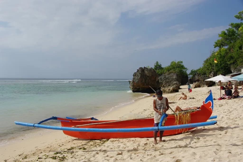 A fisherman readying a red and blue jukung outrigger on the white sand of Padang Padang Beach, Bali