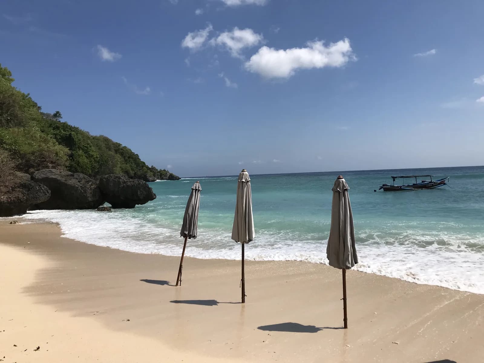 Three closed beach umbrellas on empty sand at Padang Padang in the morning, with the cliff and a boat in the bay, Bali