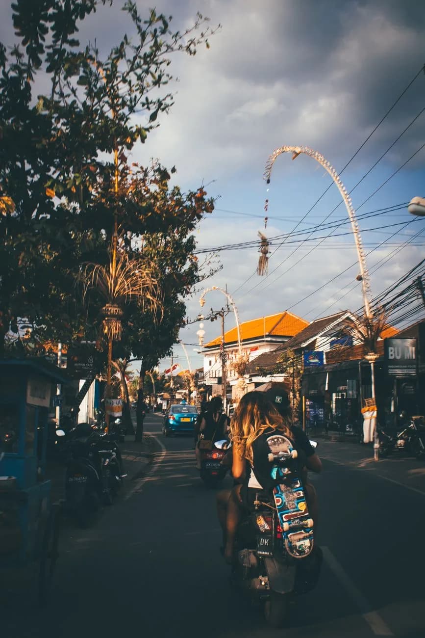 A scooter carrying a skateboard along the shop lined main street of Canggu, Bali, under ceremonial penjor poles