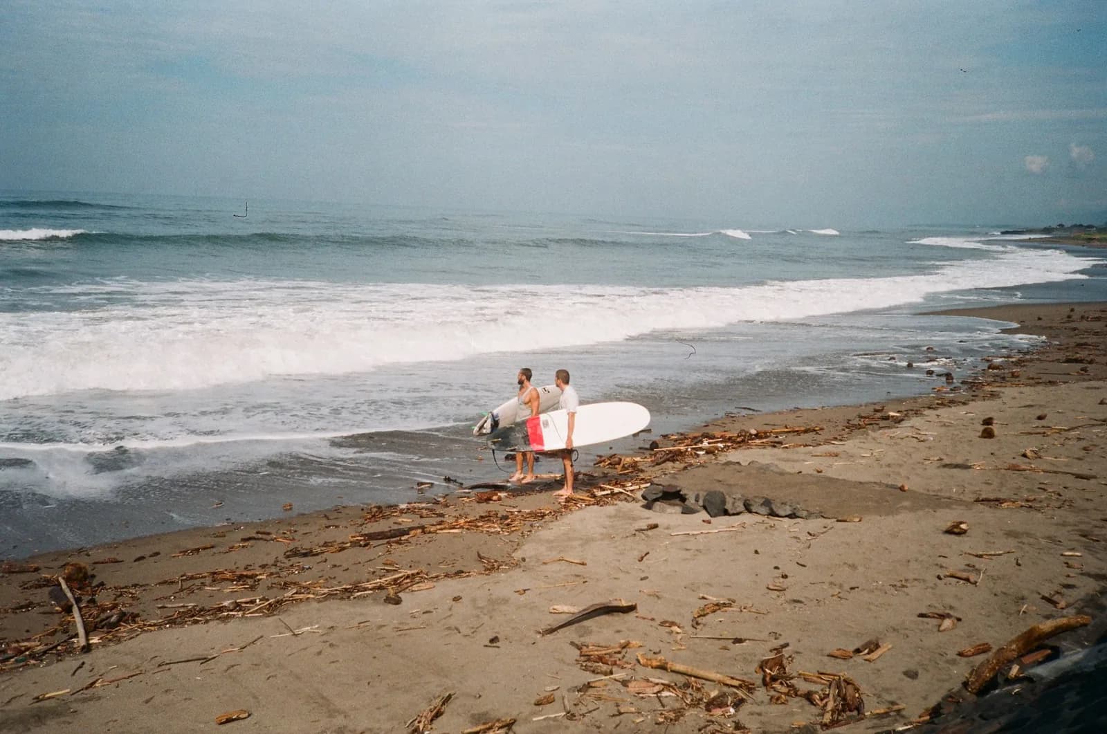 Two surfers carrying longboards across the grey sand at the waterline in Canggu, Bali