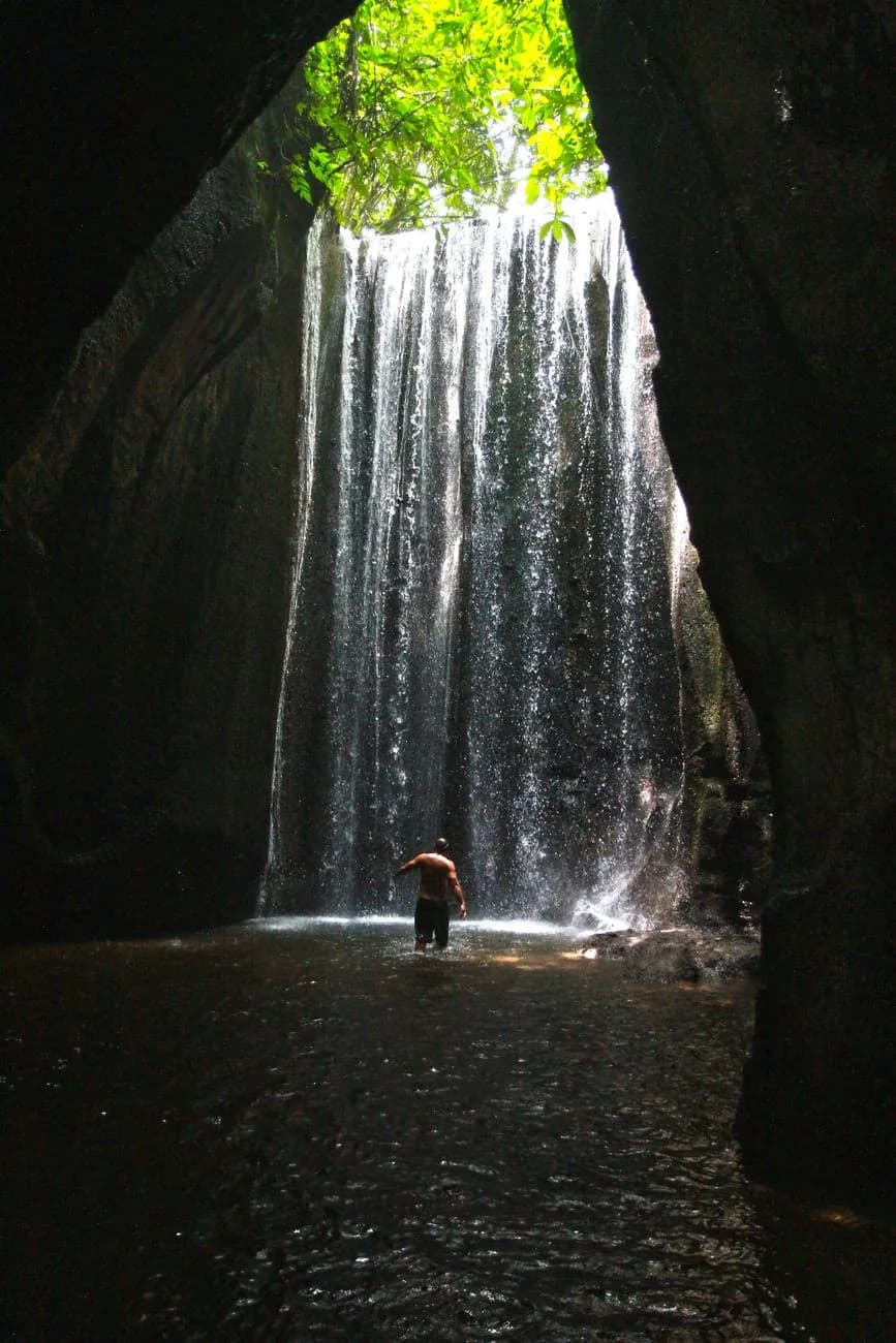 A swimmer wading through shallow dark water towards the fall inside the narrow gorge at Tukad Cepung