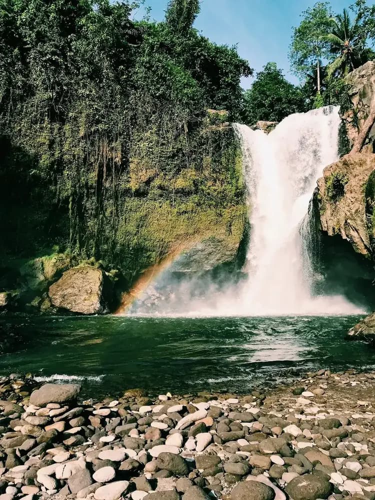 Tegenungan seen from the boulder field at river level, with a rainbow in the spray at the foot of the fall