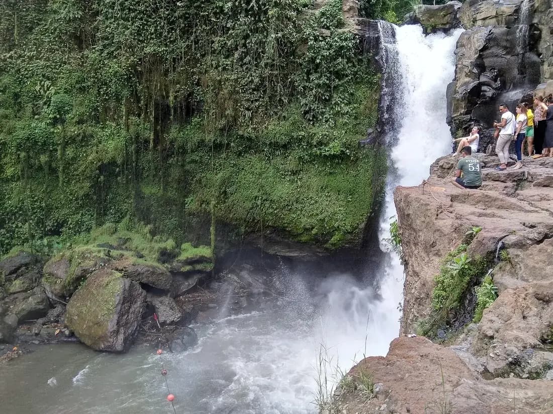 Visitors sitting on the rock ledge beside the top of the Tegenungan fall, with spray rising from the pool below