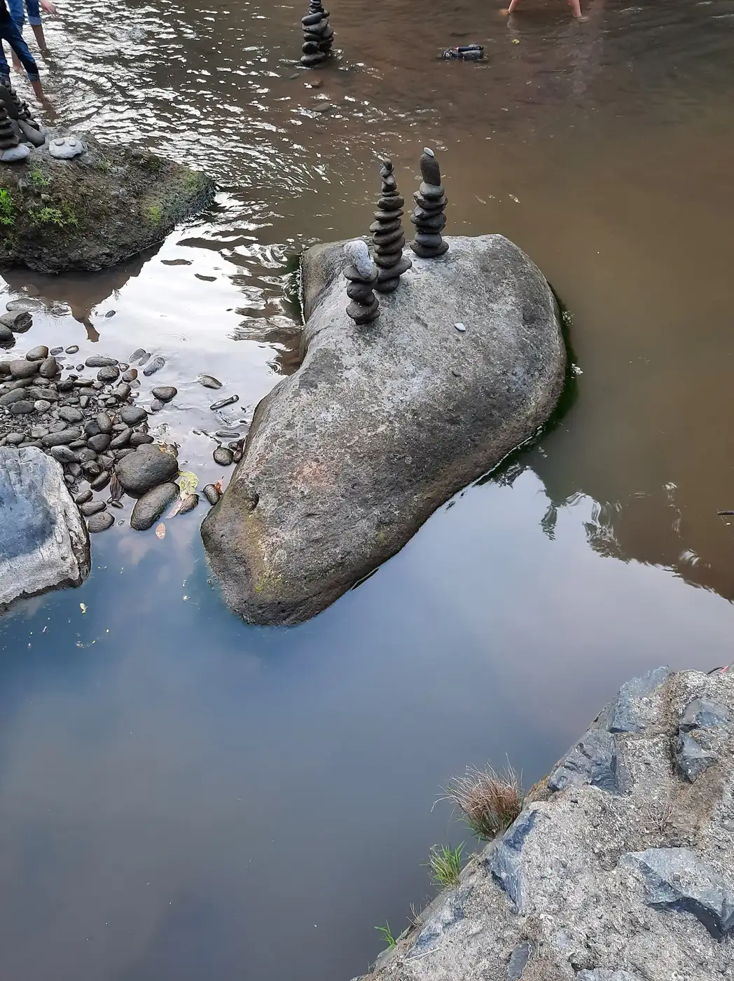 Cloudy brown river water at the base of Tegenungan with stacked stone cairns on a boulder and people wading