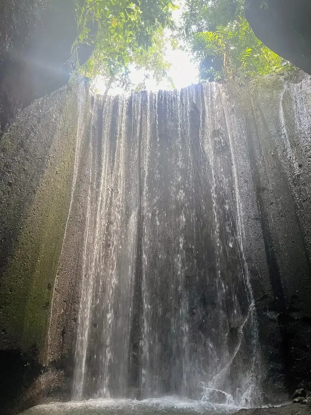 The curtain of water falling into the enclosed rock chamber at Tukad Cepung, with daylight coming through the opening above
