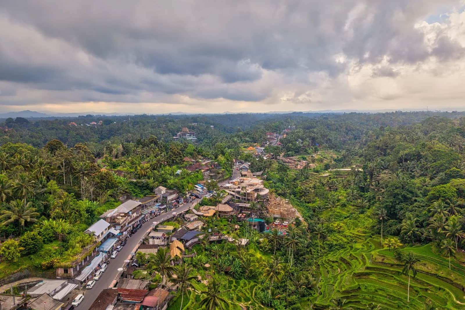 Dark monsoon storm clouds building over terraced rice fields and a café-lined road in the Bali highlands