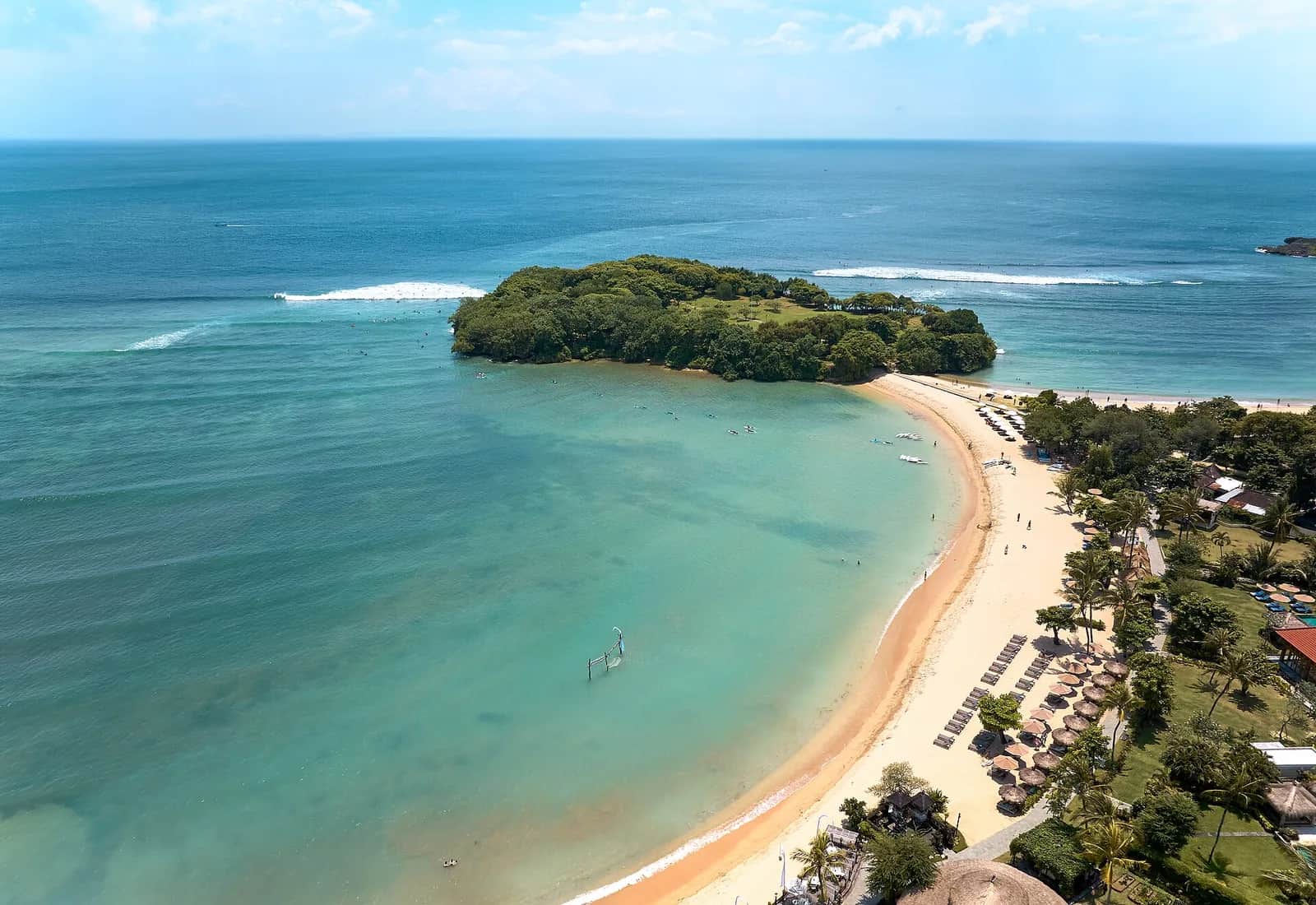 Aerial view of the calm turquoise lagoon and pale sand beach at Nusa Dua, Bali, with a small wooded islet and the reef behind