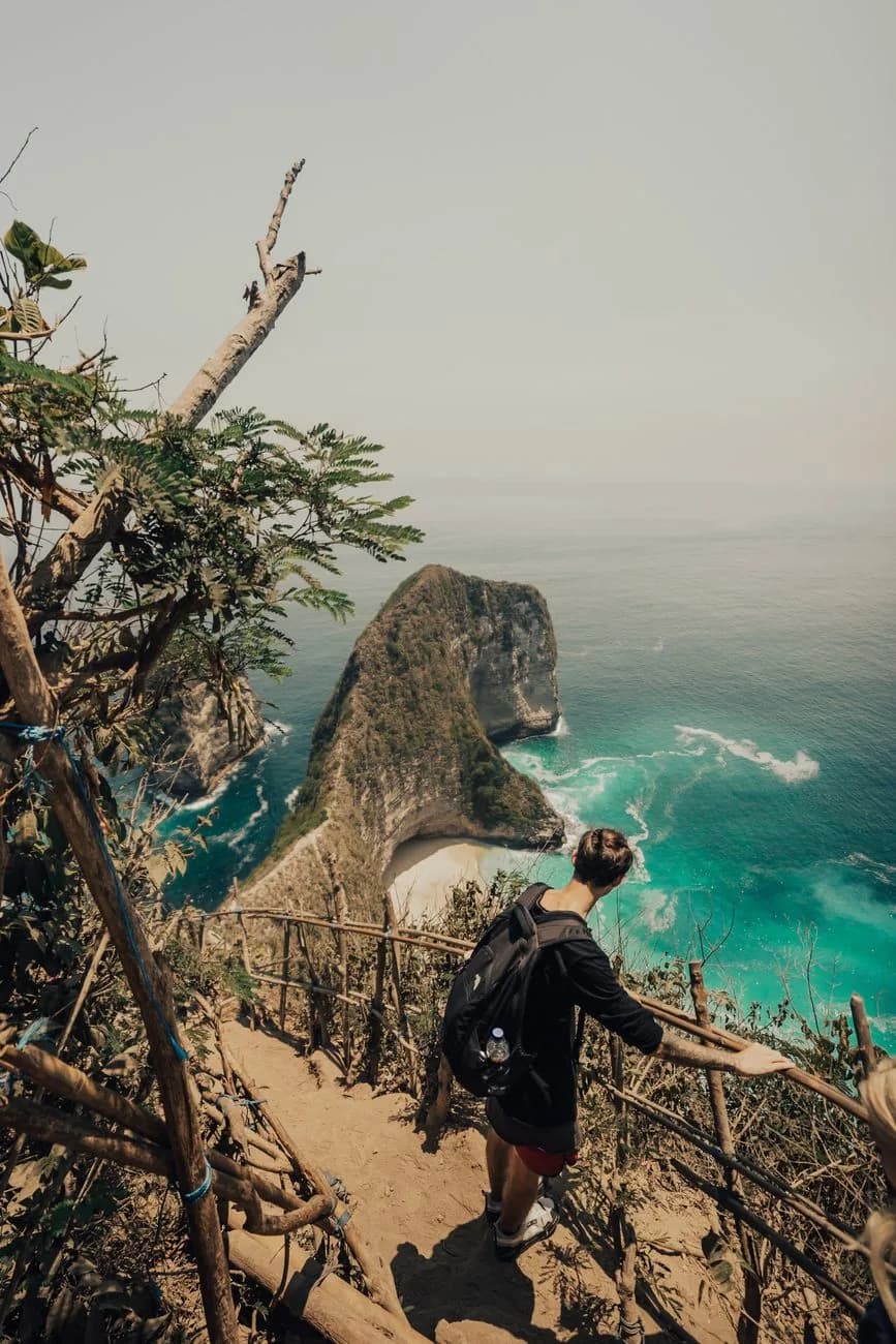 A hiker on the Kelingking Beach descent trail, holding the bamboo and branch railing above the bay, Nusa Penida