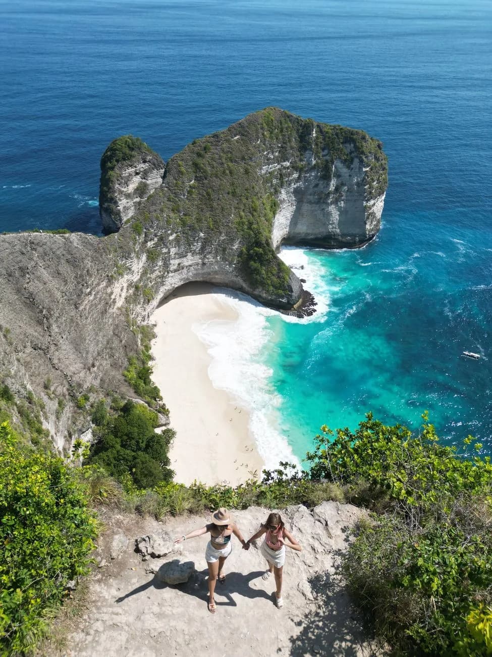 Two visitors on the rock ledge above Kelingking Beach with the headland and turquoise bay below, Nusa Penida