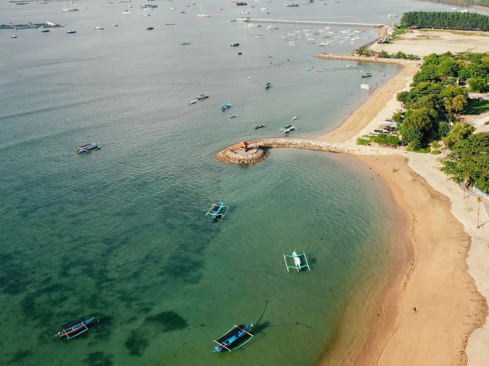 Aerial view of calm shallow water inside a reef on Bali's east coast, with traditional jukung outriggers moored off a pale sand beach