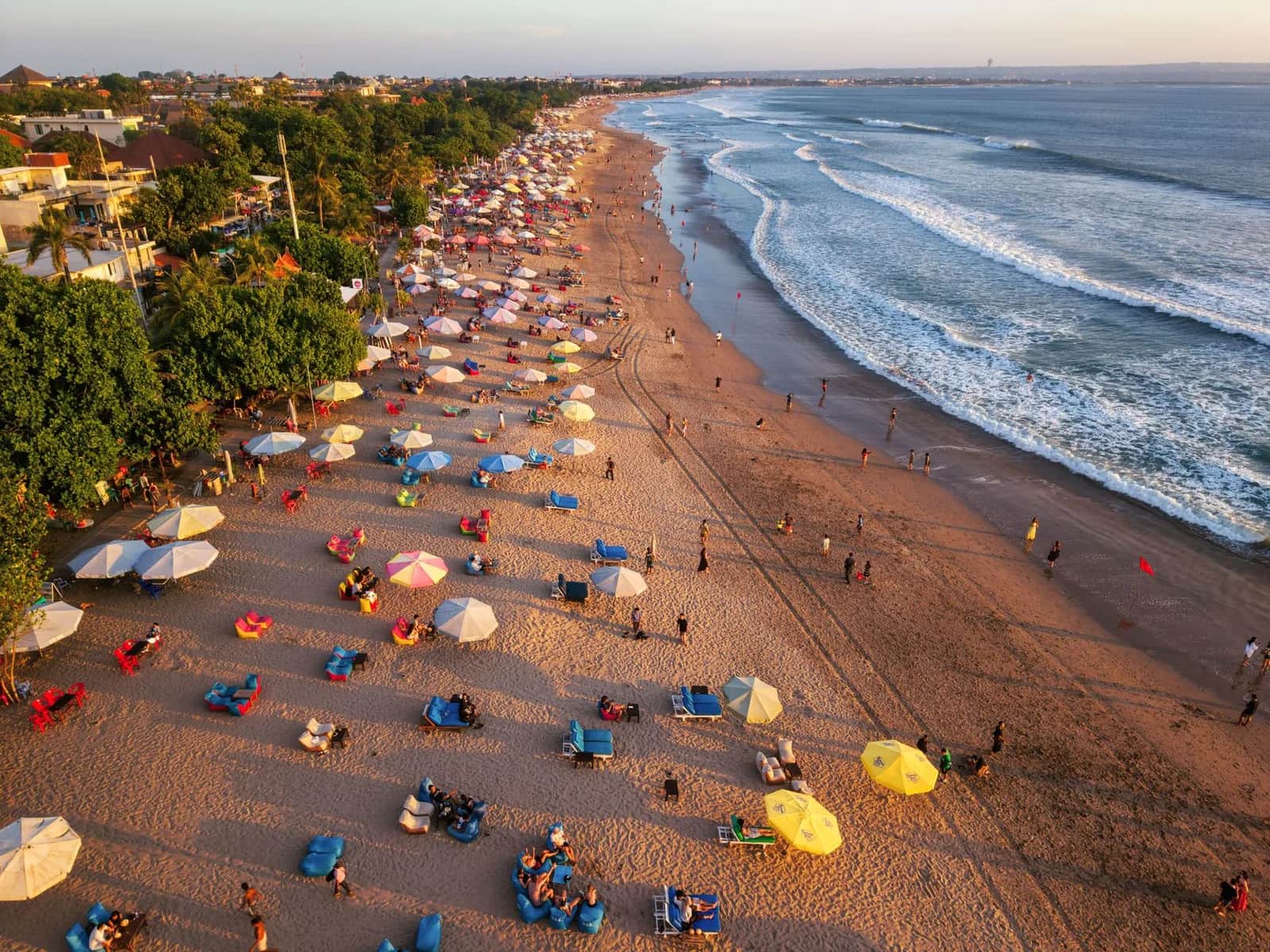 Sunset over Bali's west coast, rows of umbrellas and beanbags on grey sand, lines of surf and red flags at the waterline