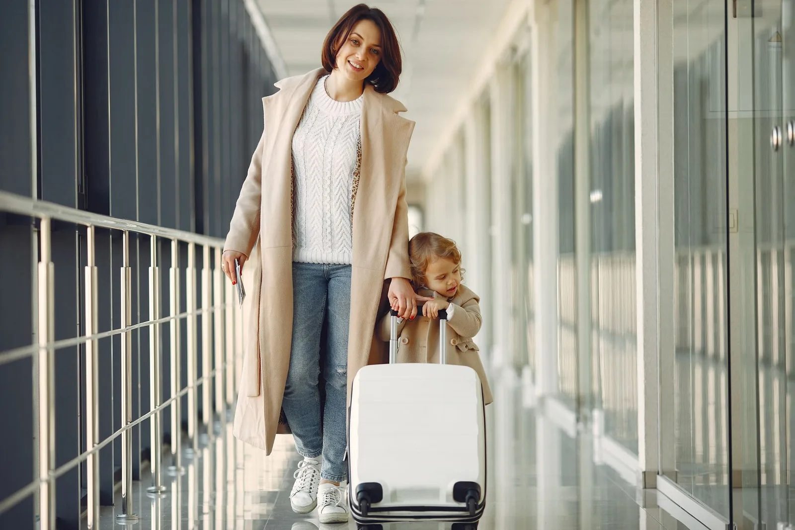 Mother and young child walking with a white suitcase through an airport corridor