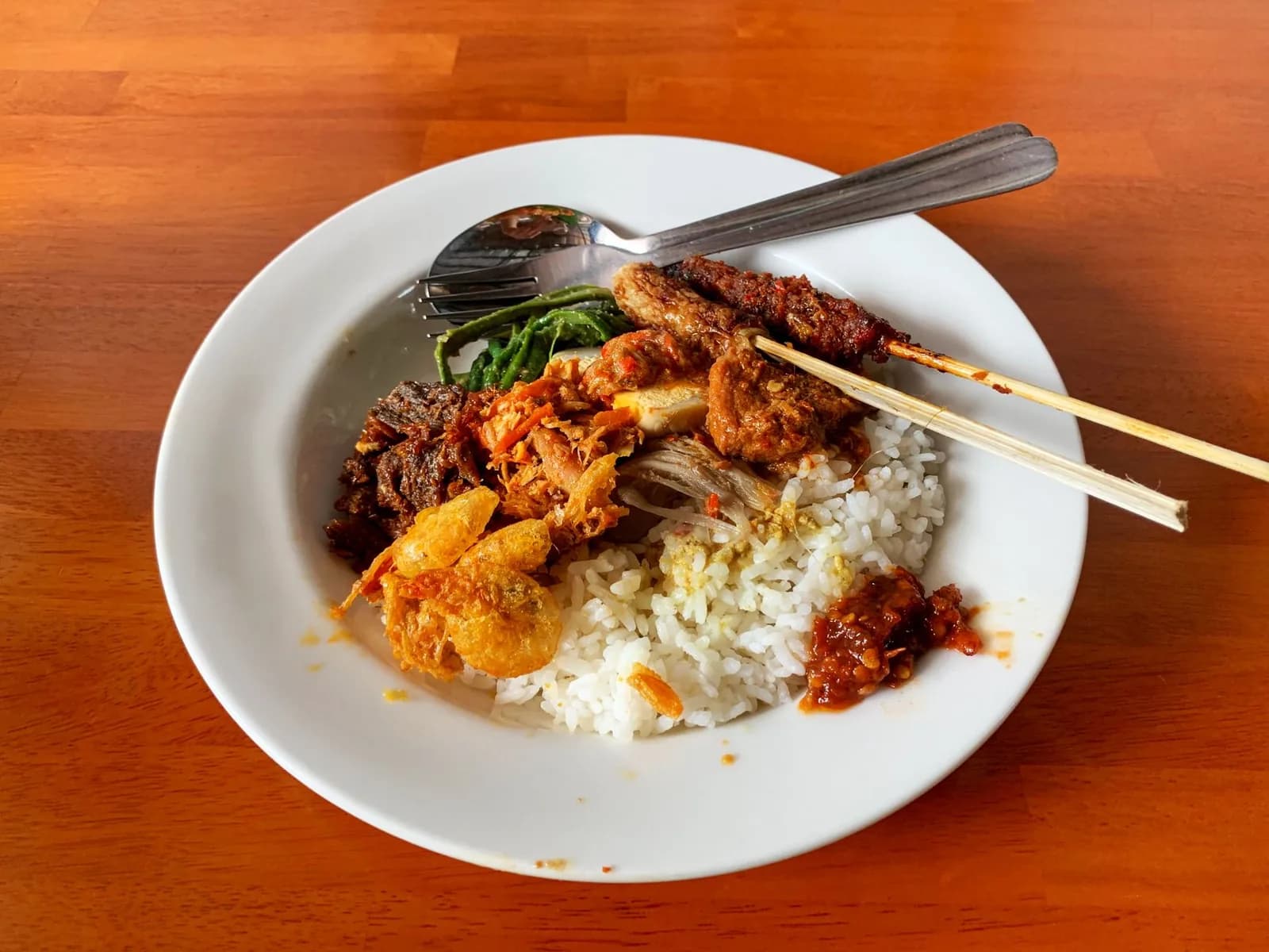 Plate of Balinese nasi campur with rice, satay, sambal and side dishes on a wooden warung table