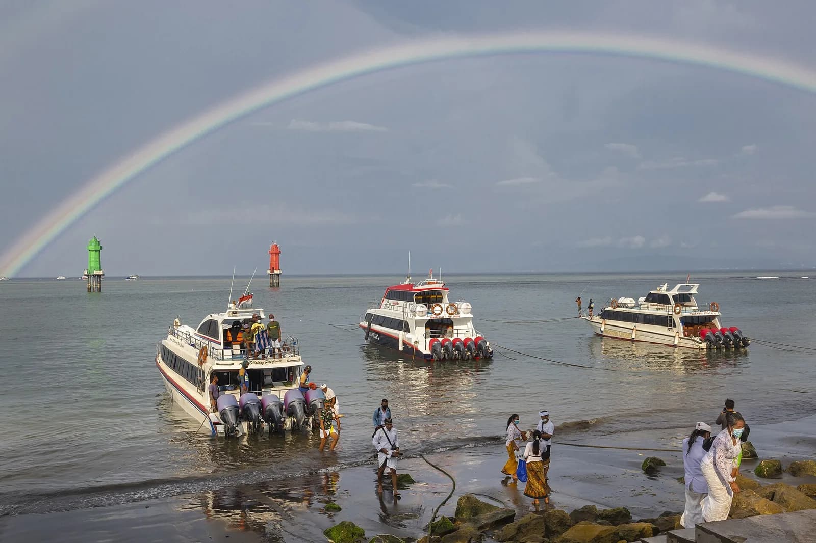 Fast boats beached at Sanur harbour with passengers boarding for Nusa Penida, under a rainbow