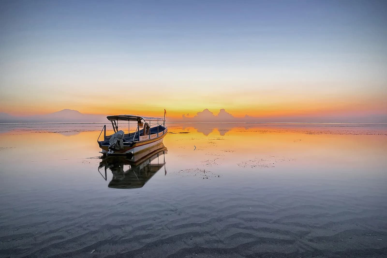 Sunrise over the still lagoon at Sanur with a fishing boat reflected in the water and Mount Agung on the horizon