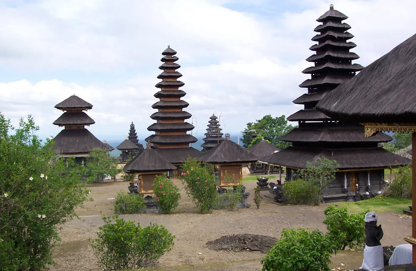 Black thatched meru towers of different heights in a Besakih courtyard, with the plain of southern Bali visible in the distance