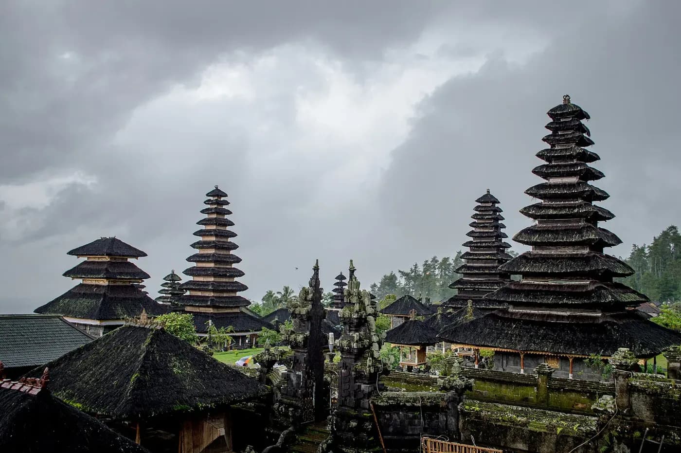 Rooflines of the Besakih temple complex under heavy grey cloud, with meru towers of eleven tiers and forest behind
