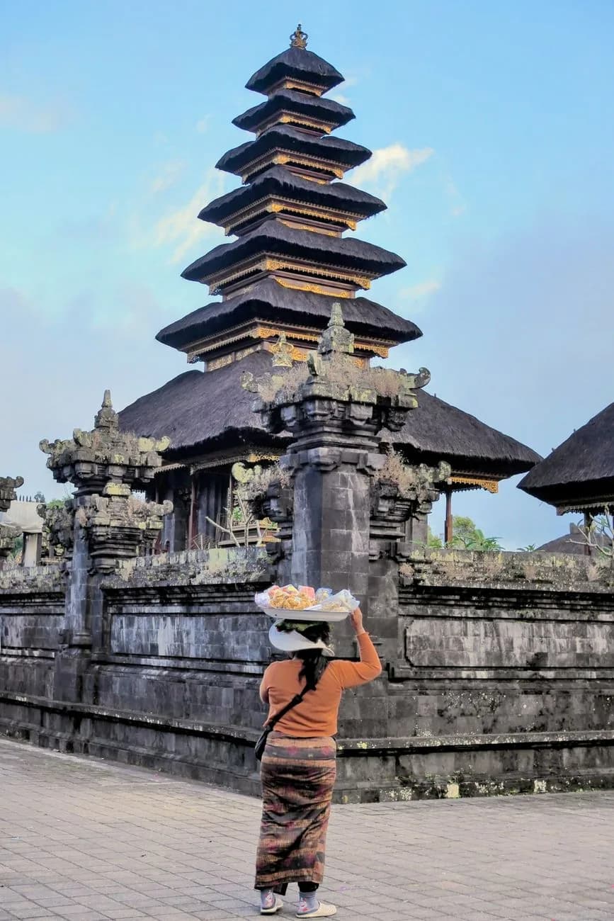 A woman carrying a tray of offerings on her head towards a meru tower at the Besakih temple complex, Bali