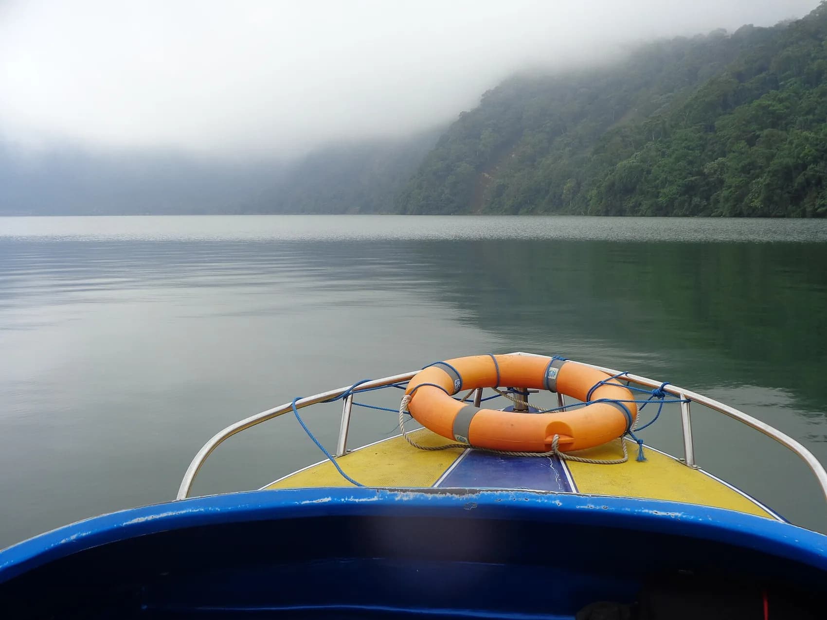 The prow of a small boat on Lake Bratan with mist covering the forested crater rim
