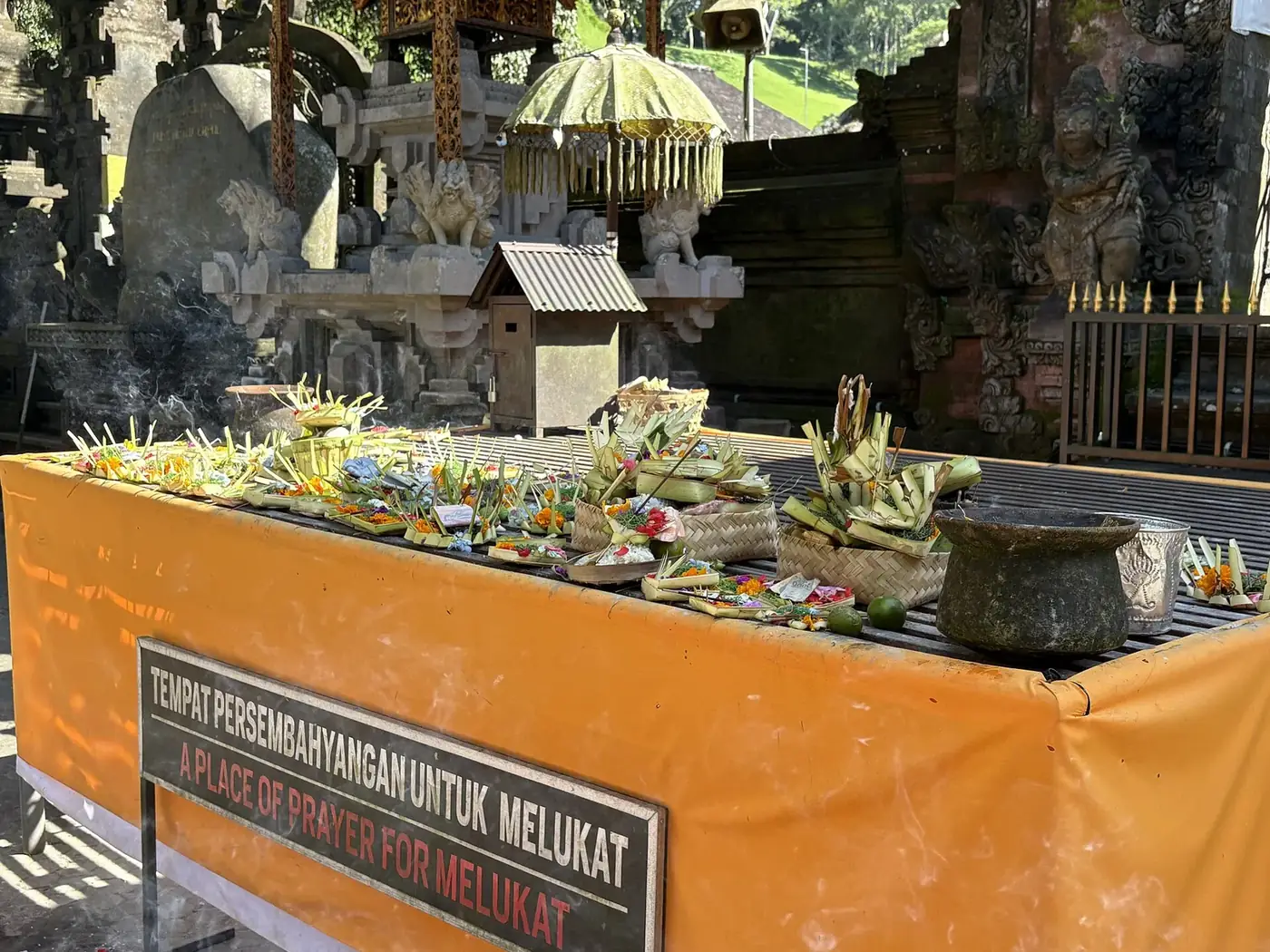 Offerings of flowers and folded palm leaf on the melukat prayer table at Tirta Empul, with an English and Indonesian sign