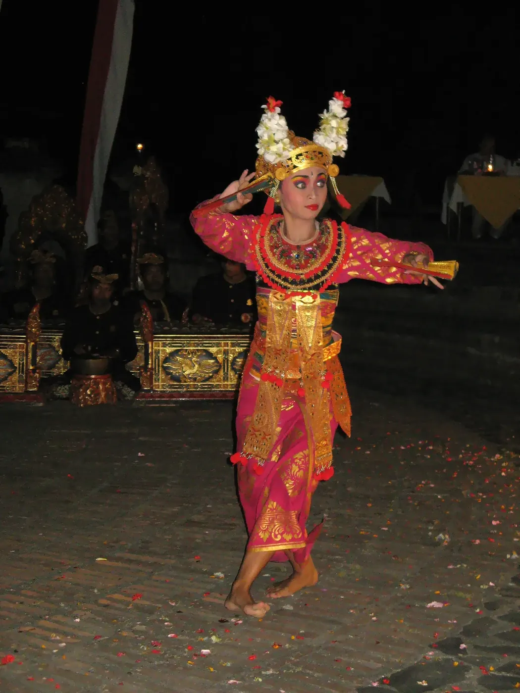 A legong dancer in pink and gold performing at night with the gamelan orchestra behind her
