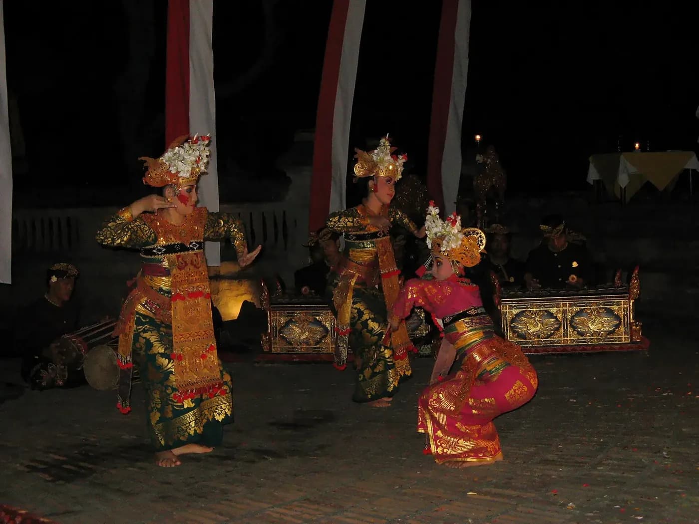 Three legong dancers in gold brocade performing at night in front of a gamelan orchestra in Ubud