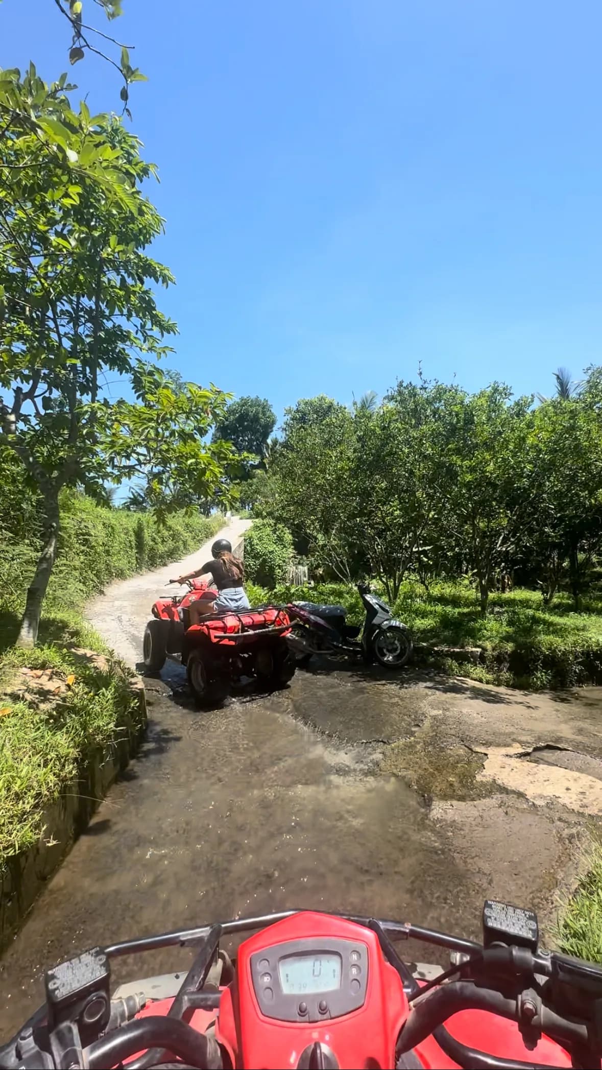 View from a quad bike's handlebars following another rider along a wet concrete track through orchards near Ubud, Bali