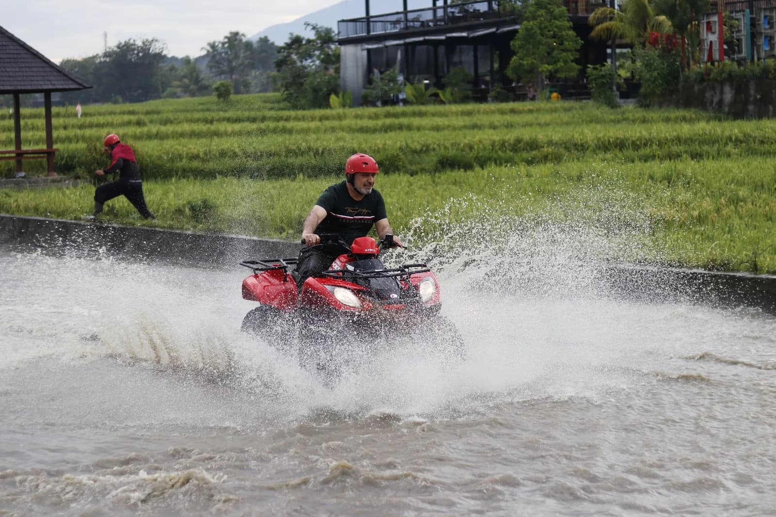 A rider in a red helmet driving a quad bike through a flooded ford beside green rice fields in Bali